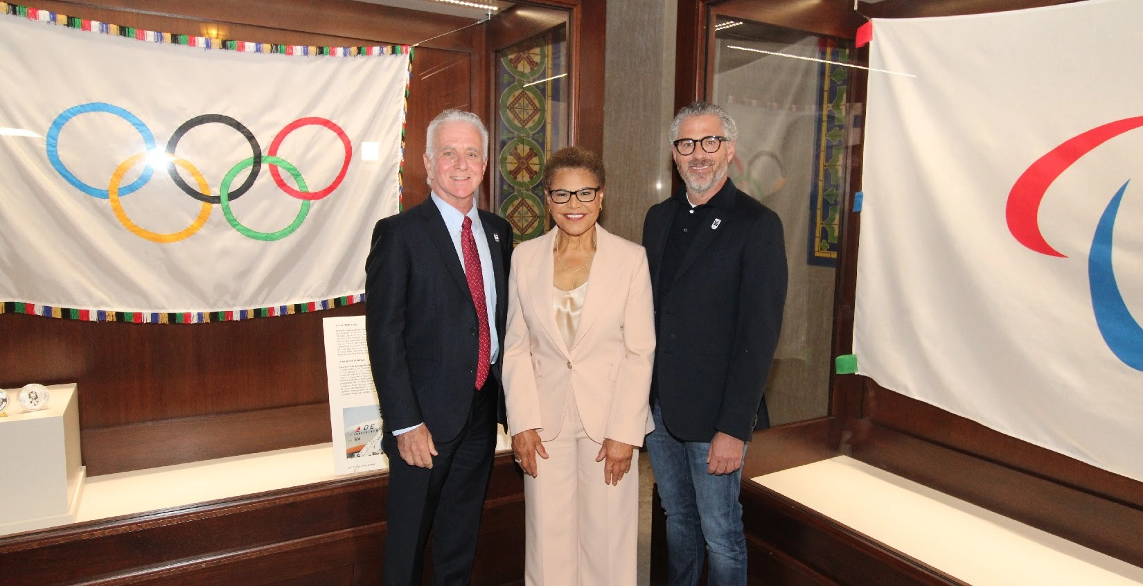 Photograph of Los Angeles Mayor Karen Bass with two men in front of displays of the Olympic and Paralympic flags
